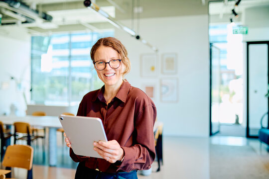 Woman standing in a bright open-plan office, holding a digital tablet and smiling at the camera, conveying professionalism and technology