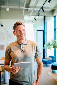 Confident man smiling, looking away while holding a digital tablet in a bright, contemporary workspace, symbolizing innovation and success