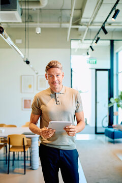 Man standing in a contemporary office, holding a digital tablet and looking at the camera, representing modern business and communication