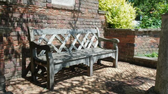 Medium shot of a weathered wooden memorial bench beside historic brickwork in a quiet shaded corner of Well Hall Park, Eltham.
