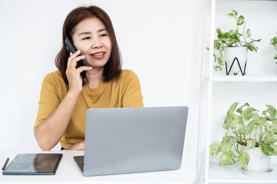 Asian business woman talking on smartphone while working on laptop in modern home office. Smiling freelancer handling customer support, telemarketing, or online consultation at desk