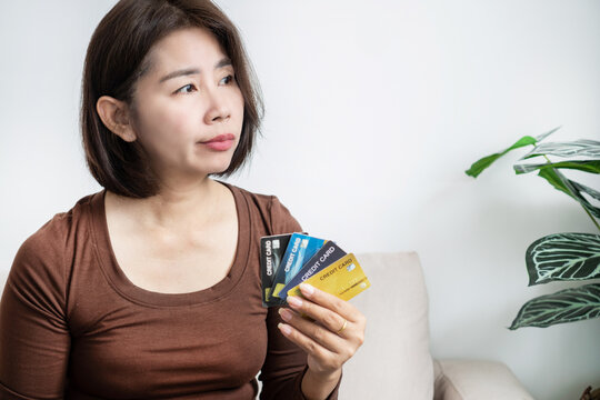 Worried Asian woman holding multiple credit cards with hesitant expression at home. Thoughtful female consumer managing personal finance, credit card debt, overspending, or payment choice