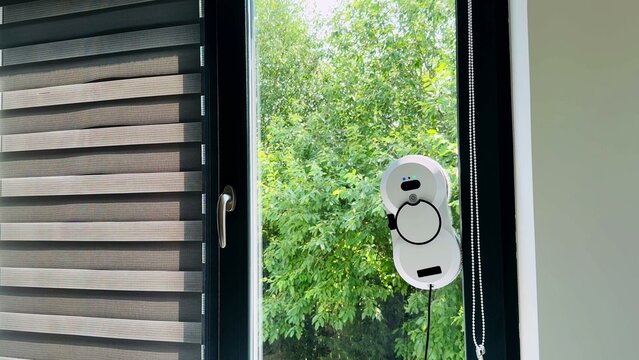 Window cleaning robot adhering to glass surface beside vertical blinds and lush green trees