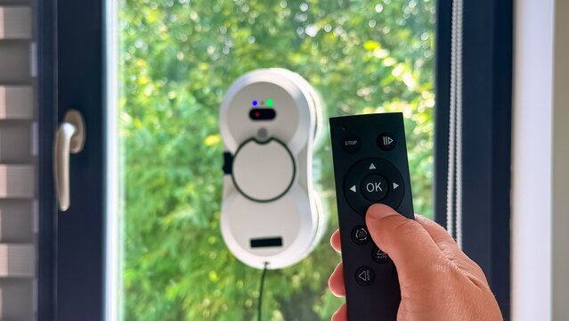 Remote control in hand activating window cleaning robot against green foliage background