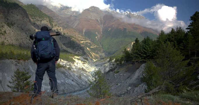 Annapurna Circuit, Manang, Nepal, camera shot of a man with a backpack stepping into the frame to look over the Marsyangdi River flowing through a deep valley beneath Himalayan mountain peaks.