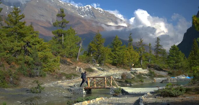 Annapurna Circuit, Manang, Nepal, camera shot of a hiker standing on a small wooden bridge along an alpine trekking trail surrounded by pine forests and high mountain peaks.