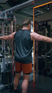 Muscular man seen from behind raises arms sideways training side deltoids on cable machine in dark gym - vertical shot