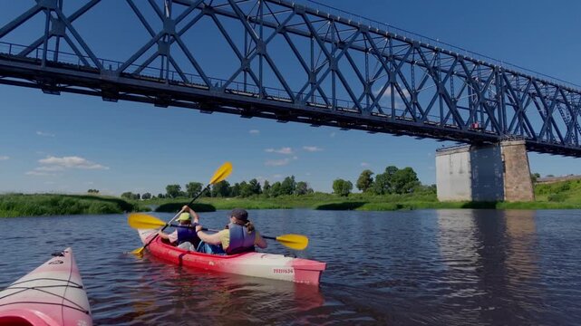 Active tourists paddle tandem red kayak along wide river under massive steel railway truss bridge structure. Sunny summer recreation activity in beautiful Baltic region.