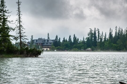 A tranquil view of &Scaron;trbsk&eacute; Pleso lake in the High Tatras, Slovakia, with morning mist drifting above the water and a mountain hotel surrounded by evergreen forest. Dramatic clouds, calm waters, and lu