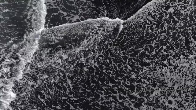 Top down aerial view of white ocean foam waves crashing and washing over black sand beach shore