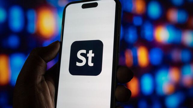 Smartphone showing adobe stock icon on screen held by hand against colorful glowing digital backdrop.