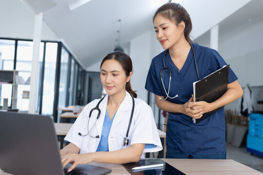 Two smiling Asian female healthcare professionals, a doctor and a nurse, discussing medical reports together in a modern hospital office.	
