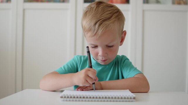 Focused little boy writing in notebook in school. Small child scribbling with a pen in a book. Child focused on practicing handwriting in school. Boy learning to write in kindergarten.