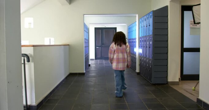 African American school-age girl walking down school hallway past blue lockers toward double doors