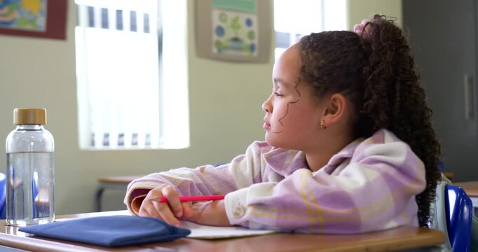 Elementary-school girl watching then pausing, writing to complete worksheet with red pencil at desk