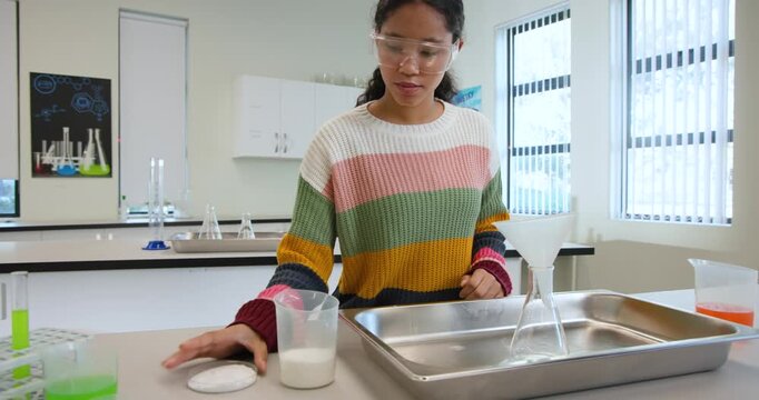 Asian child securing goggles, pouring white liquid via funnel into flask filtering at lab bench