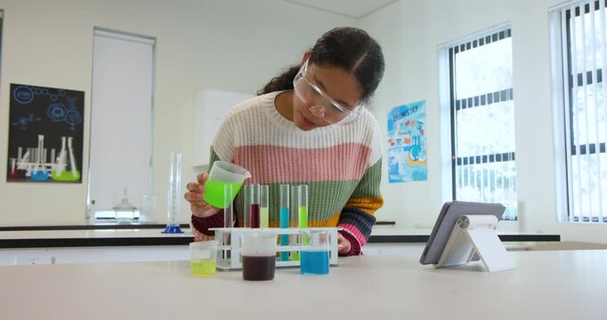 Following tablet Asian child wearing goggles pouring bright liquids into test tubes at school lab