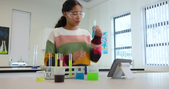 Female child in goggles, sweater holding test tube, inspecting color at lab bench following tablet