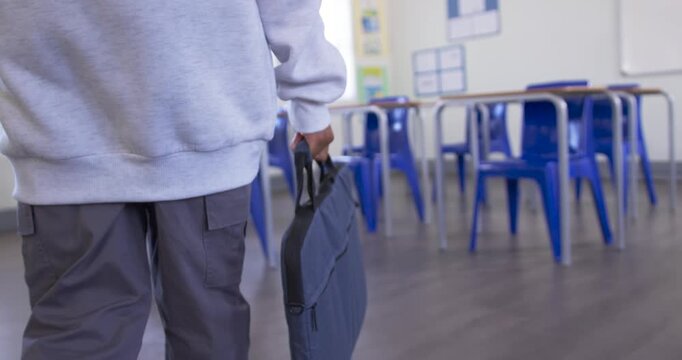 Teenage male walking through class carrying laptop bag, adjusting grip past blue chairs, copy space