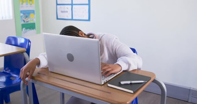 African American teen lifting head from laptop tugging headphones typing for class at school desk