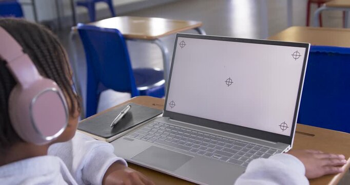 African male youth sitting left at school desk wearing pink headset studying on laptop, copy space