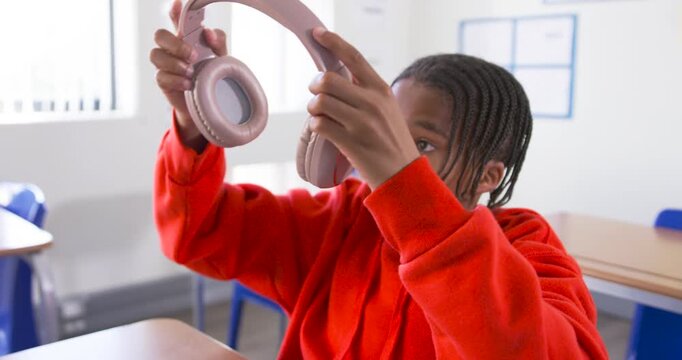 African teen in red hoodie lifting pink headphones on ears, inspecting fit, falling asleep at desk