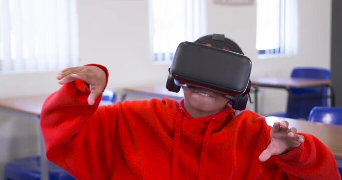 African American teen in red hoodie reaching for tablet at desk, adjusting VR headset during class