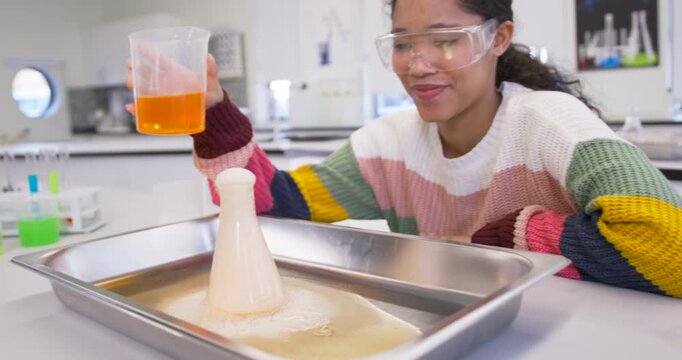Female child in goggles pouring orange liquid from beaker into flask on lab tray showing eruption