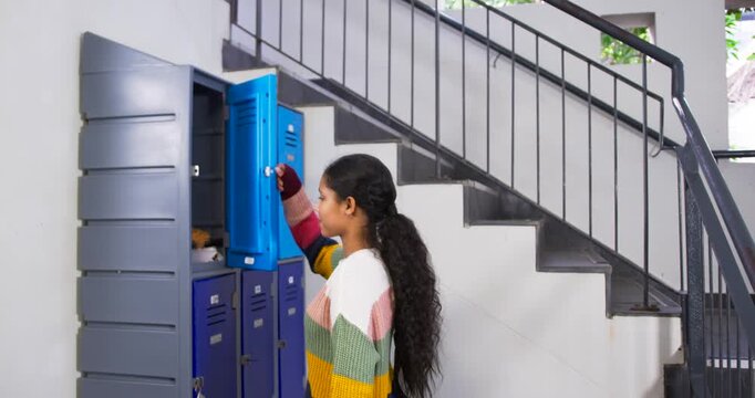 Girl needing bag at lockers by stairs, using handheld, unlocking top blue locker, pulling out bag