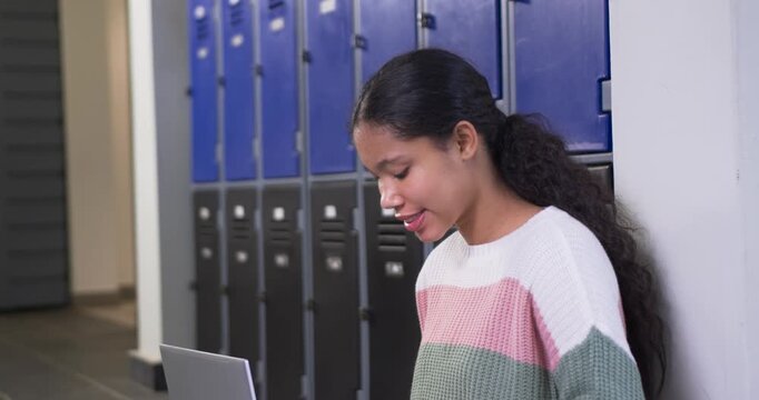 Female child sitting against pale wall in school hall beside lockers, smiling then typing on laptop