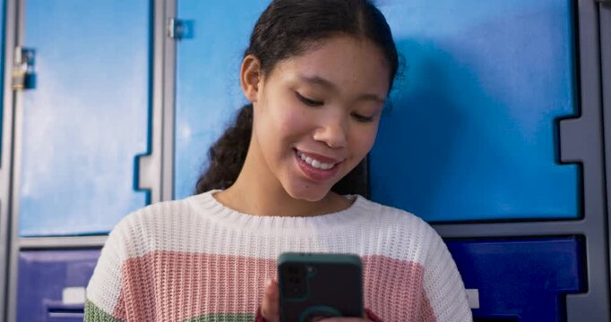 Asian child girl leaning against blue school lockers in knit sweater, checking phone with grip