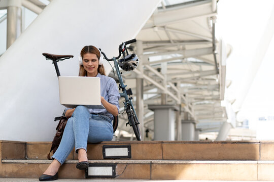 Young Woman with Laptop and Bicycle: A young woman in casual attire sits on steps with her laptop and a cycle beside her, suggesting bike to work and an active lifestyle in an urban setting.