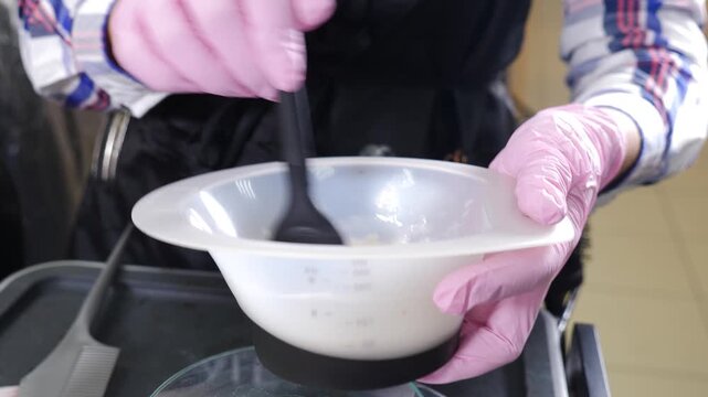 Close-up of hairdresser pink-gloved hands mixing hair dye in clear plastic bowl using black spatula. Professional salon preparation process before coloring treatment, precise work and clean tools. 4k