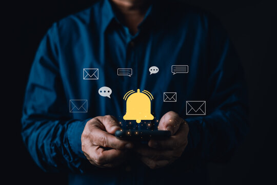 Man holding smartphone with glowing yellow notification bell, email, and chat icons. Concept of digital communication, new messages, social media alert, and online reminder on dark background.