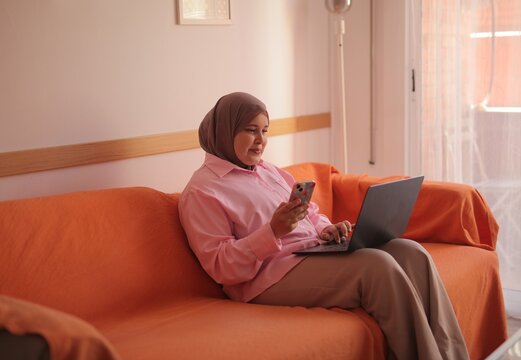 Smiling muslim woman multitasks on phone and laptop