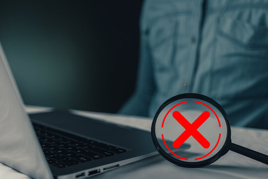 Concept of rejection, error, and problem. Laptop and magnifying glass with red X sign showing denial, fail, incorrect, delete, or prohibit message. Dark background with blurred person.