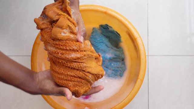Person washing dirty orange towel in basin with soapy water alongside blue clothes