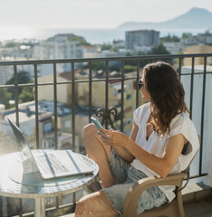 Obraz premium Millennial woman holding smartphone with open laptop in front of her on sunny balcony with city view.