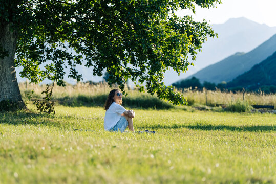 Beautiful young woman relaxing on green grass under a large tree, enjoying summer nature.