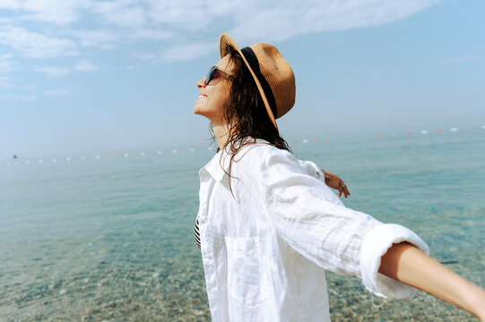 Happy young woman in summer hat and white shirt standing on sea beach, enjoying freedom with arms stretched back.