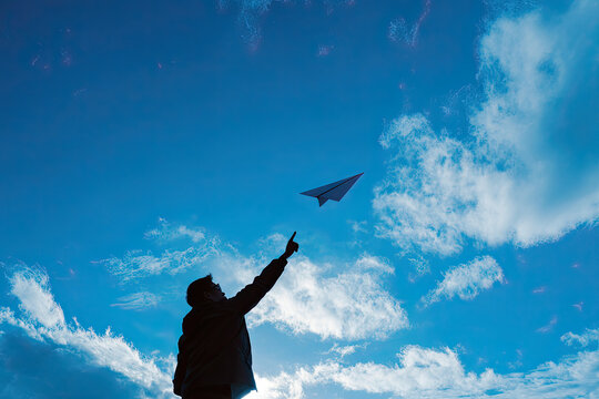 Silhouette of passionate young man throws folded paper airplane toward beautifully bright cloudy blue sky, expressing deep desire for total freedom, creative startup ideas, and genuine hope