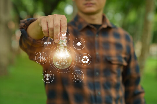 Man holding glowing light bulb with clean energy icons compared to unlit bulb in green nature background.