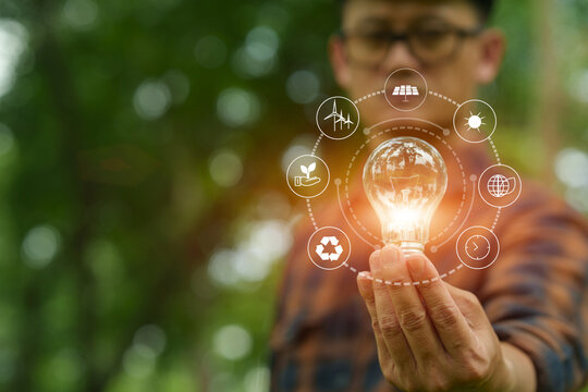 Man holding glowing light bulb with clean energy icons compared to unlit bulb in green nature background.