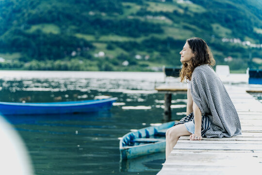 Side view of peaceful young woman wrapped in blanket sitting on wooden pier and looking at mountain lake.