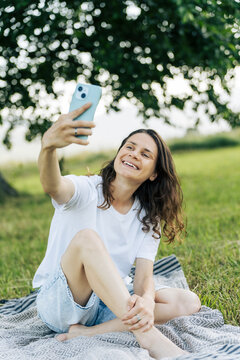 Happy young woman sitting on picnic blanket and taking a selfie with smartphone under a large tree.