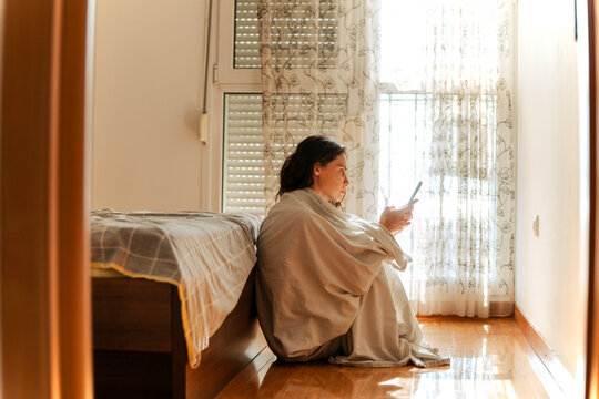 Focused young woman wrapped in white sheet sitting on floor by bed and looking at smartphone.