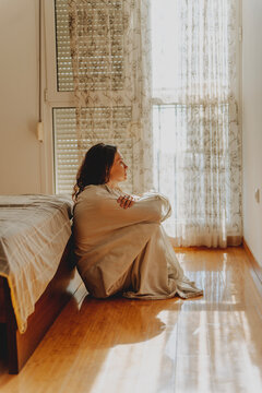 Thoughtful young woman wrapped in white sheet sitting on floor by bed, hugging knees with neutral expression.