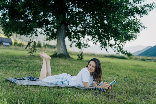 Happy young woman lying on stomach on picnic blanket and using smartphone on green grass.