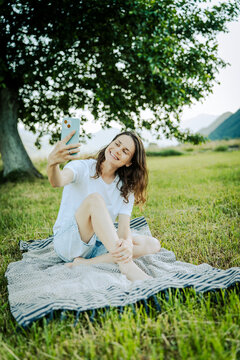 Happy young woman sitting on picnic blanket and taking a selfie with smartphone under a large tree.