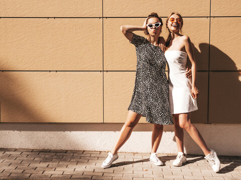 Two young beautiful smiling hipster in trendy summer dresses. Sexy carefree women posing in the street near yellow wall.Positive models having fun in sunglasses